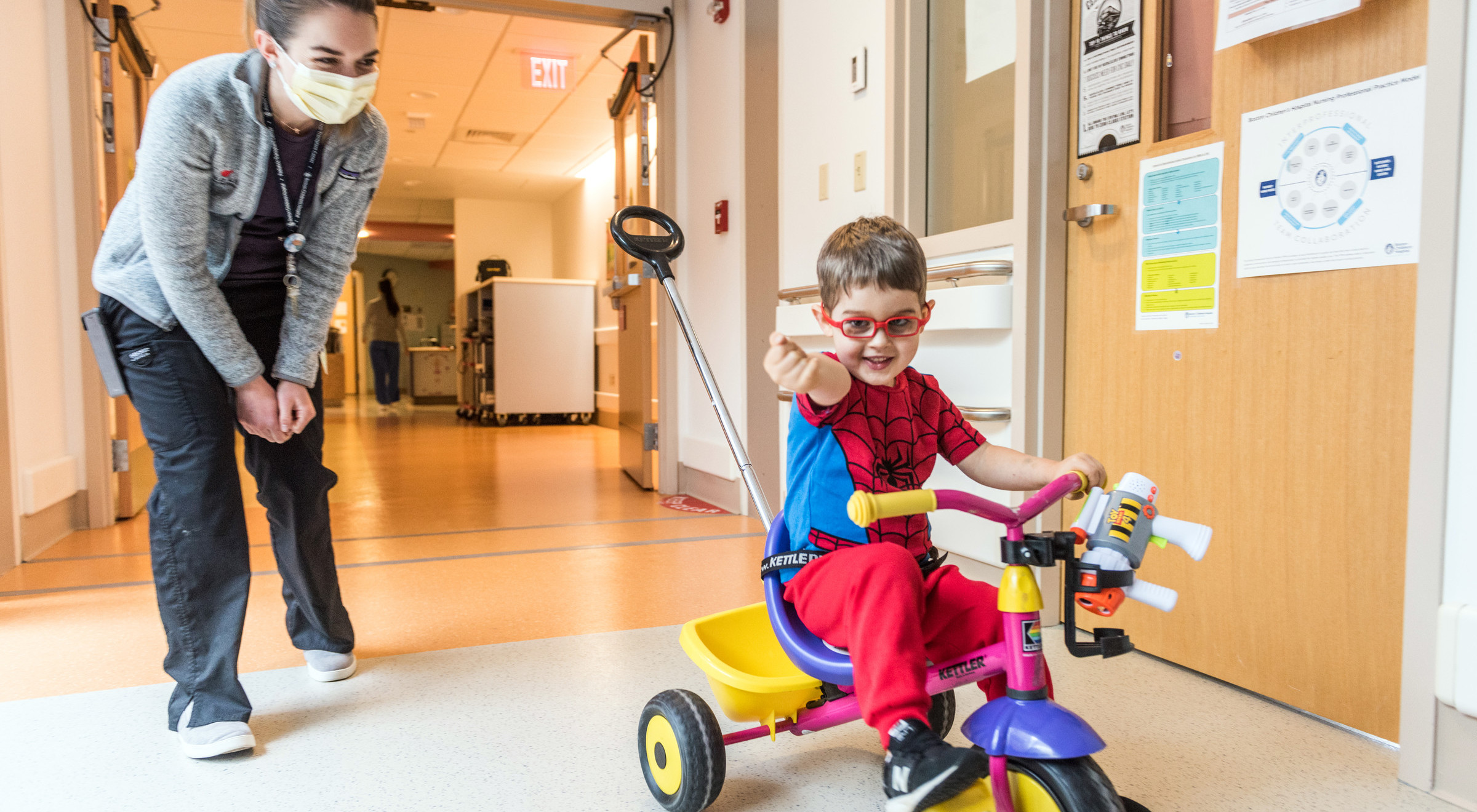 Boy rides tricycle through hallways of medical unit while watched by a clinician.