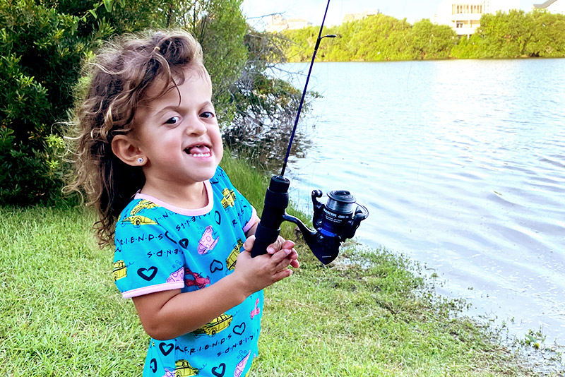 Young girl wearing blue shirt holds fishing rod in her hands while standing alongside water