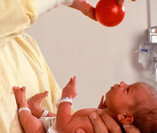 A doctor holds a small red ball over a newborn baby.