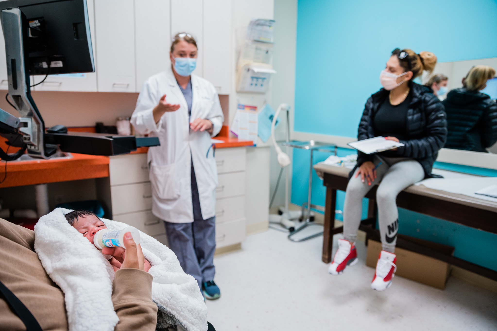 a swaddled newborn feeding from a bottle while a pediatrician talks to a parent