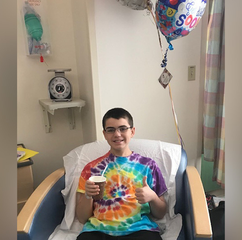 A boy in a tie-dye t-shirt sits in a hospital bed and gives a thumbs up sign.