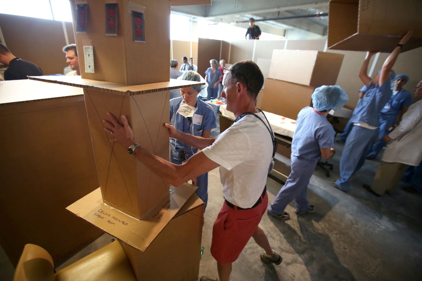 Doctors in blue scrubs and a man in red shorts and a white shirt all move cardboard boxes around in a room as they use them to build a building.