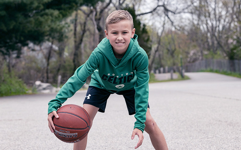 Boy wearing Boston Celtics green sweatshirt dribbles ball on roadway