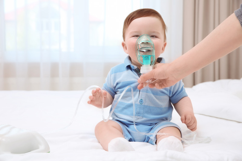 baby on a bed with a hand holding a breathing device to his face