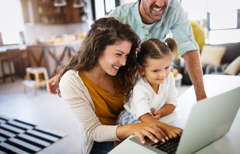 a daughter sits on her mom's lap while looking at a computer together and a man is behind them