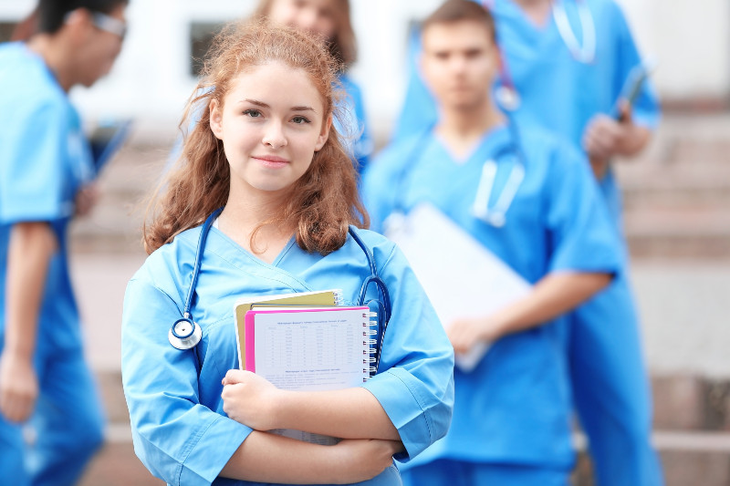 a pretty young woman with a stethoscope around her neck holding notebooks and looking at the camera