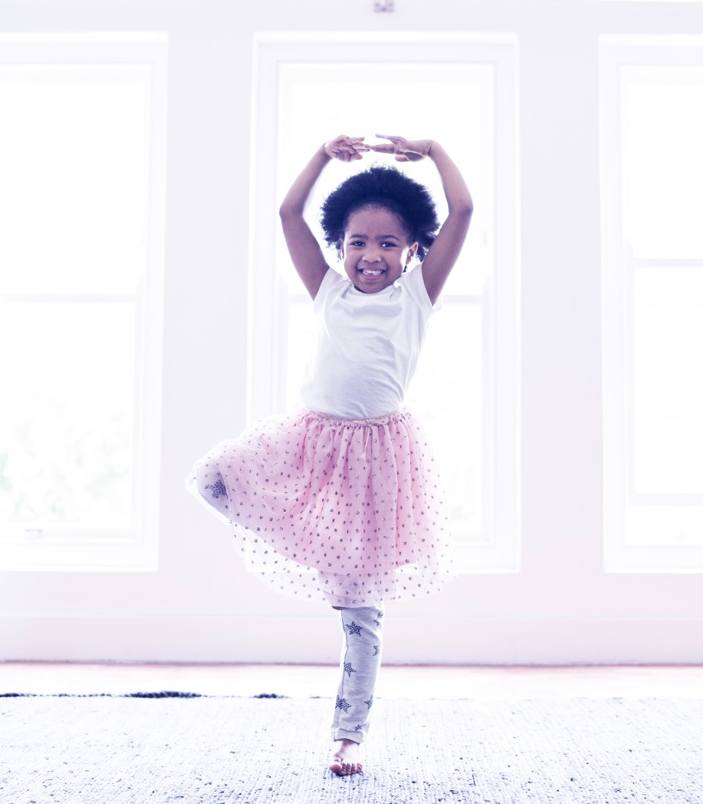 A black girl standing in white shirt and pink skirt stands in front of windows in her ballerina pose. 