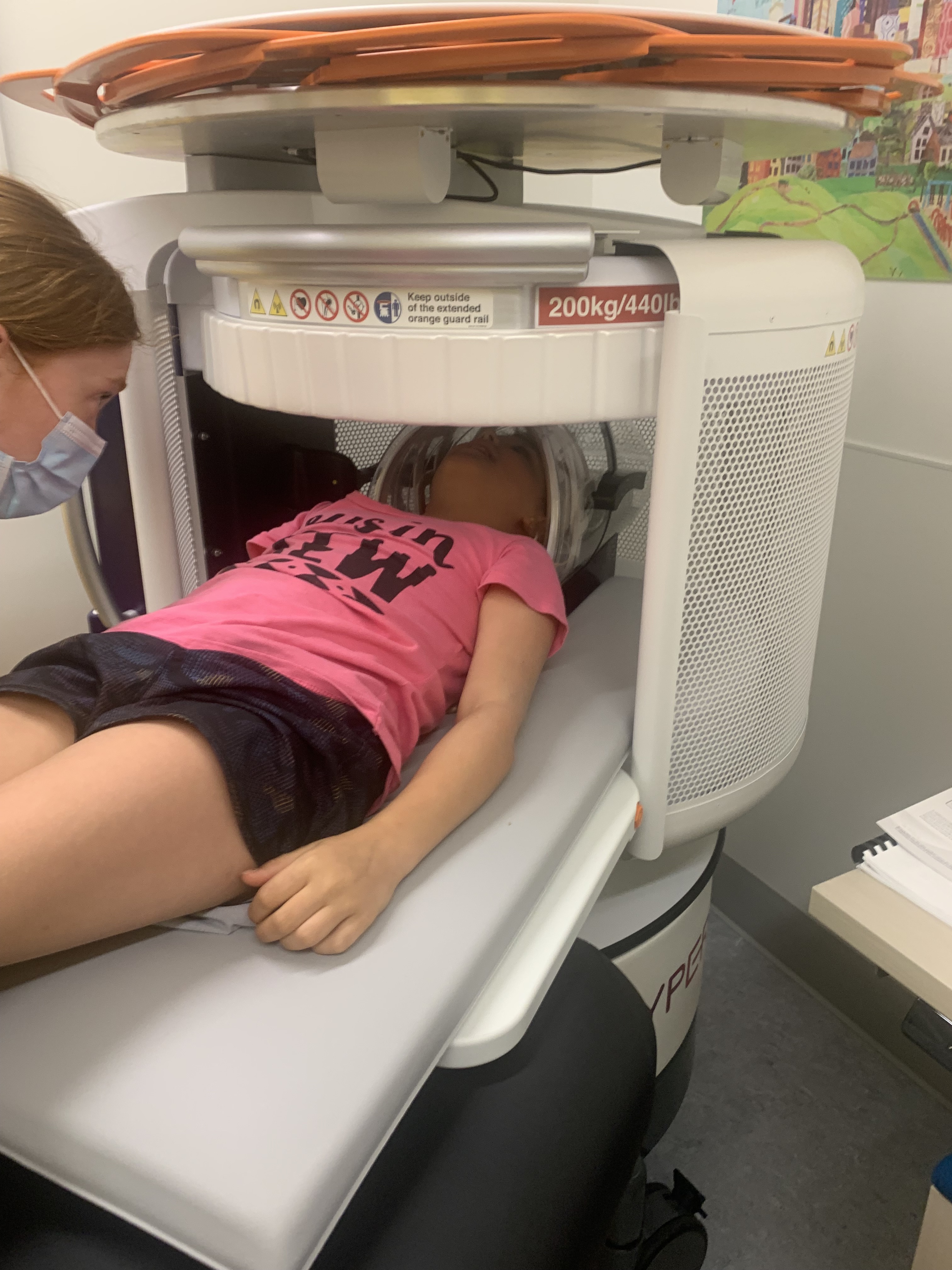 A girl in a pink shirt and black shorts lays in an MRI machine that scans her brain.