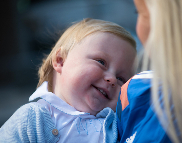 Smiling boy with blonde hair held by woman.