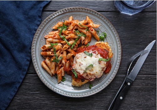 Baked chicken parmesan with pasta on the side on a silver plate with a fork and knife on the table next to the dish.