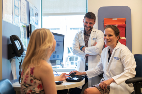 Two doctors facing a blond woman around a table in a room that looks like a doctors office.