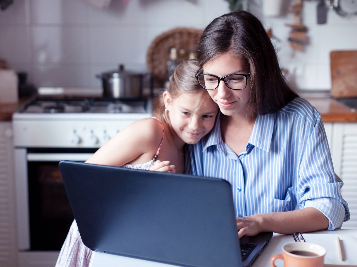 A mother with glasses works on her computer while her daughter leans in to her.