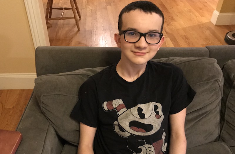 Young boy wearing glasses and black T-shirt sits on gray couch