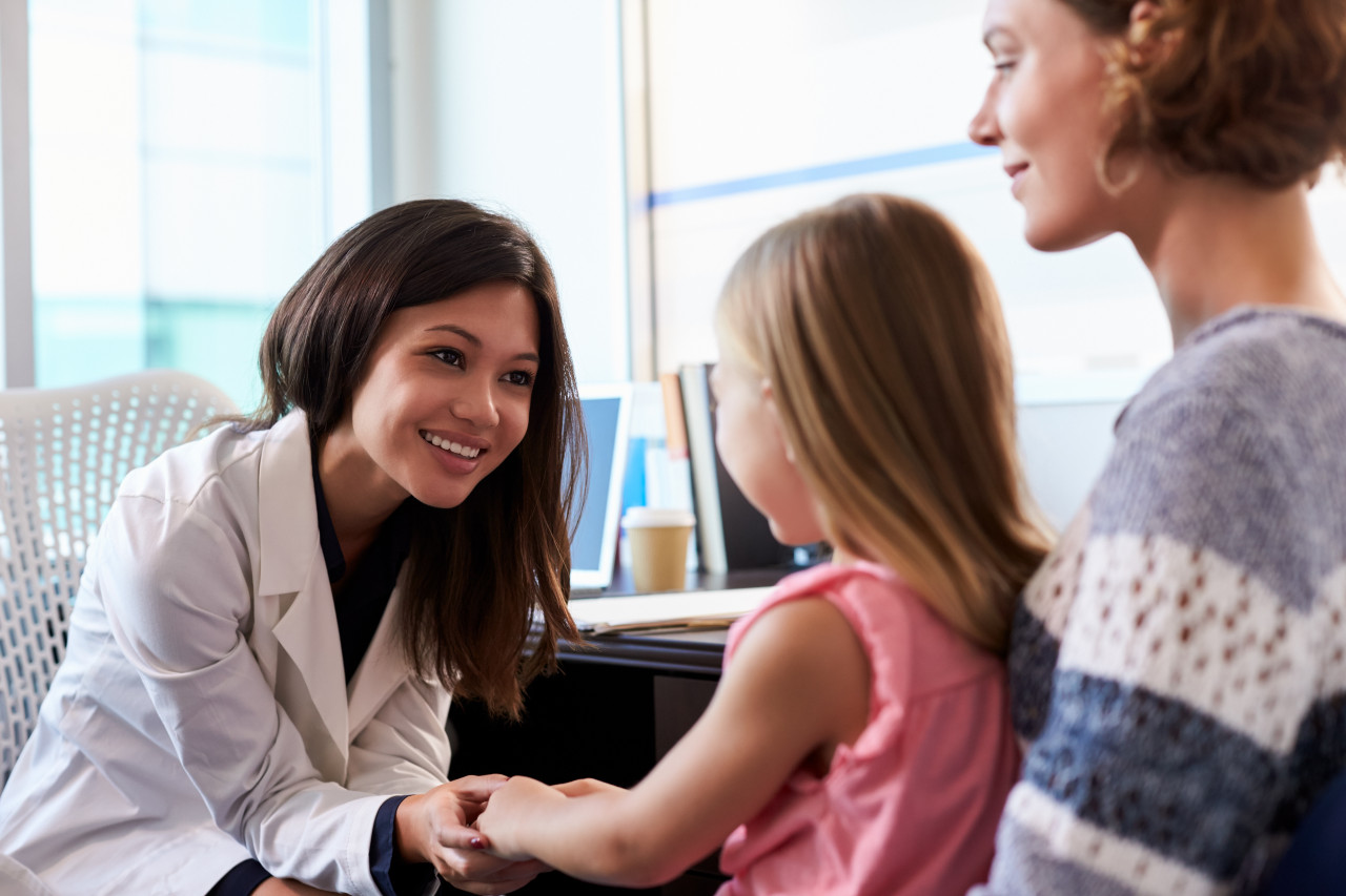 a woman pediatrician smiling at a young girl who is sitting on her mom's lap