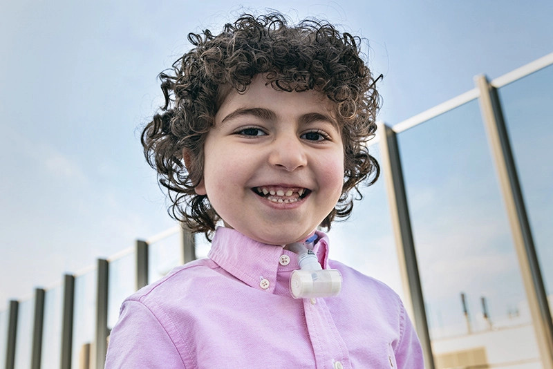 Young boy with curly hair and wearing a pink shirt smiles while standing in front of a fence