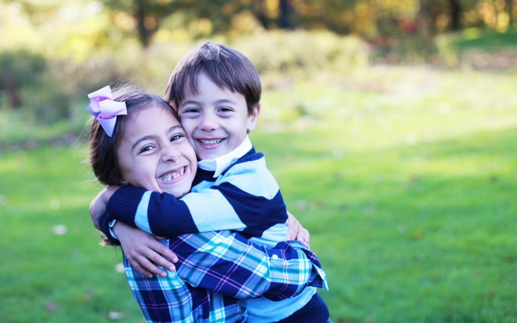 Young boy in striped rugby shirt hugs sister wearing flannel shirt and bow in hair