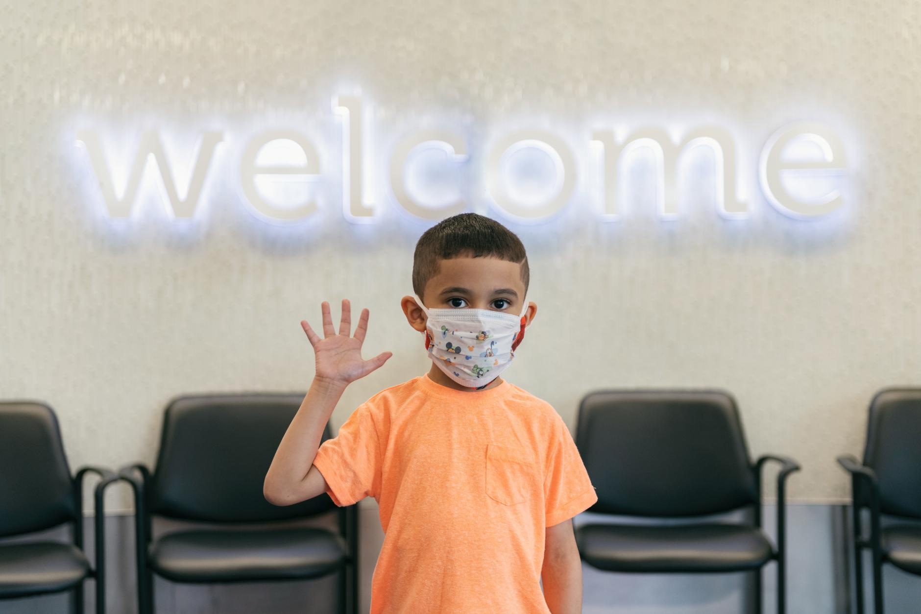 Boy wearing face mask waives at camera while standing in front of "welcome" sign
