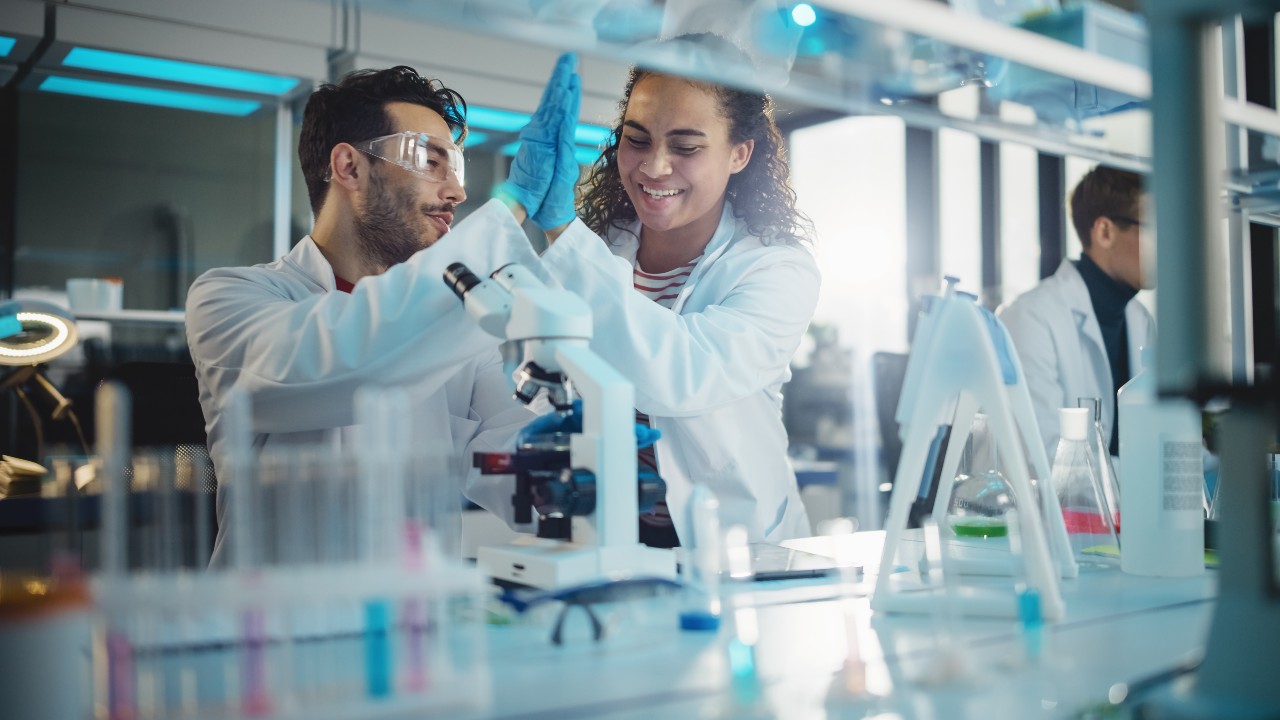 Two researchers high five each other behind a table filled with test tubes and a microscope.