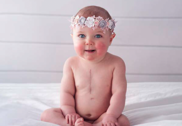 Smiling baby, with red cheeks and wearing a crown of flowers, looks at the camera.