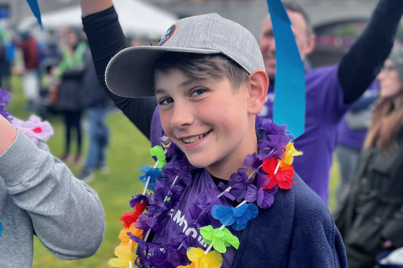 Boy wearing purple shirt, gray hat and lei smiles for camera
