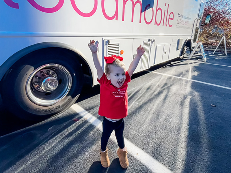 Young girl stands in front of the Boston Children's Bloodmobile, with arms raised