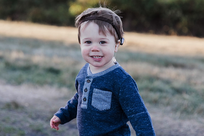 Small boy wearing blue long-sleeve shirt and bone-anchored hearing system smiles for photo