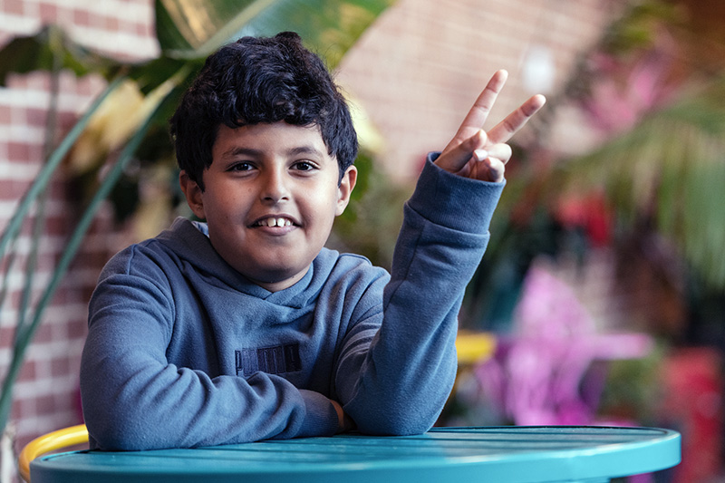 Boy wearing blue sweatshirt gives "peace" sign to camera.