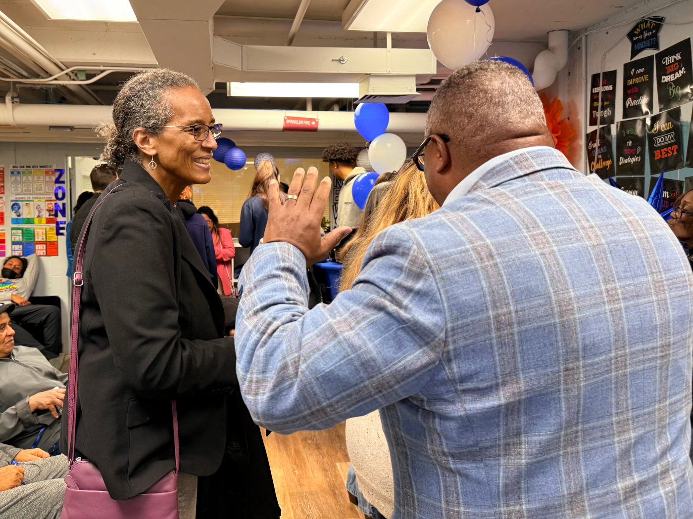 Man wearing glasses, with back to camera, speaks to woman wearing glasses.