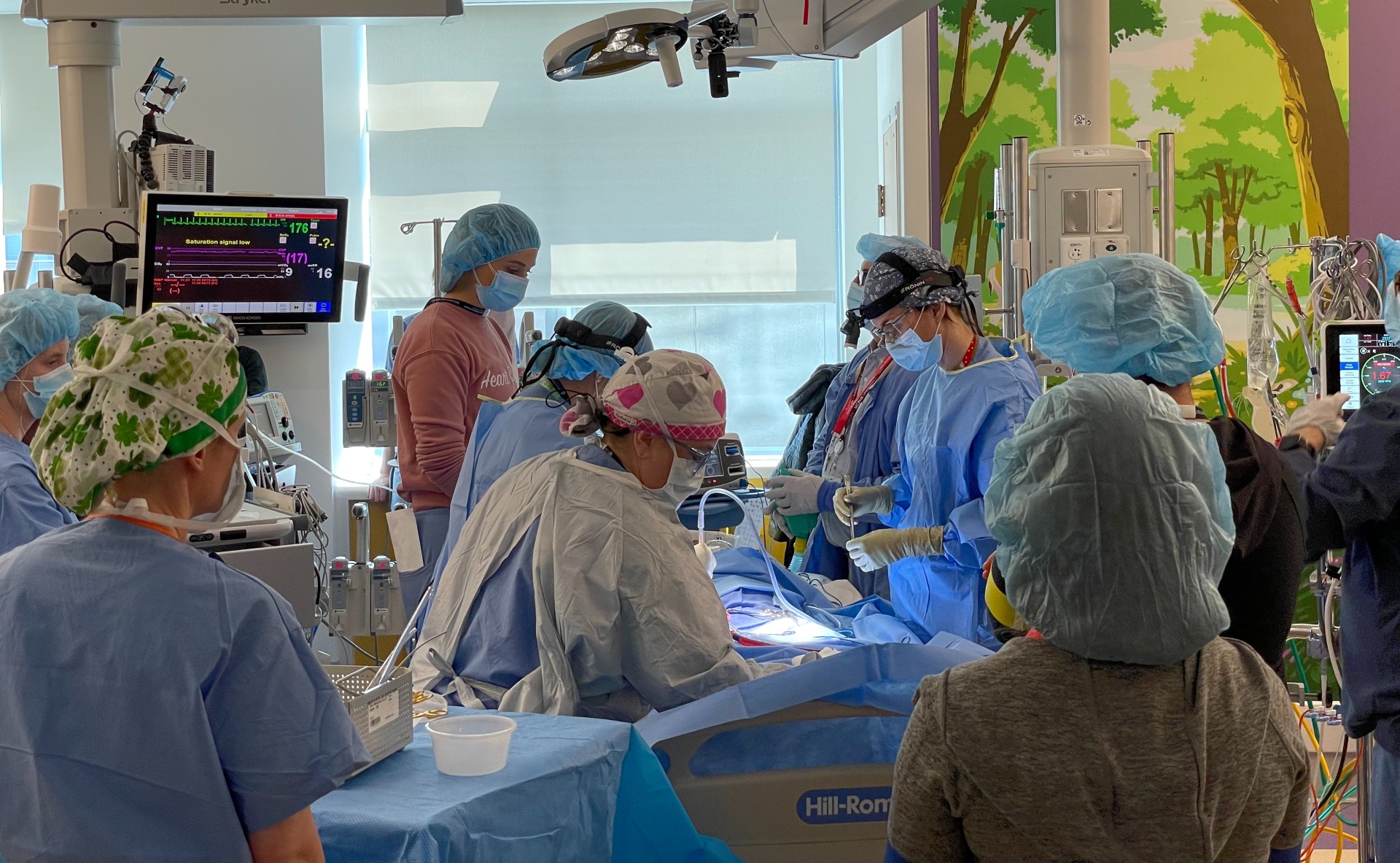 A group of doctors in blue scrubs perform on a simulation child in a room with a pink wall and a tree mural.