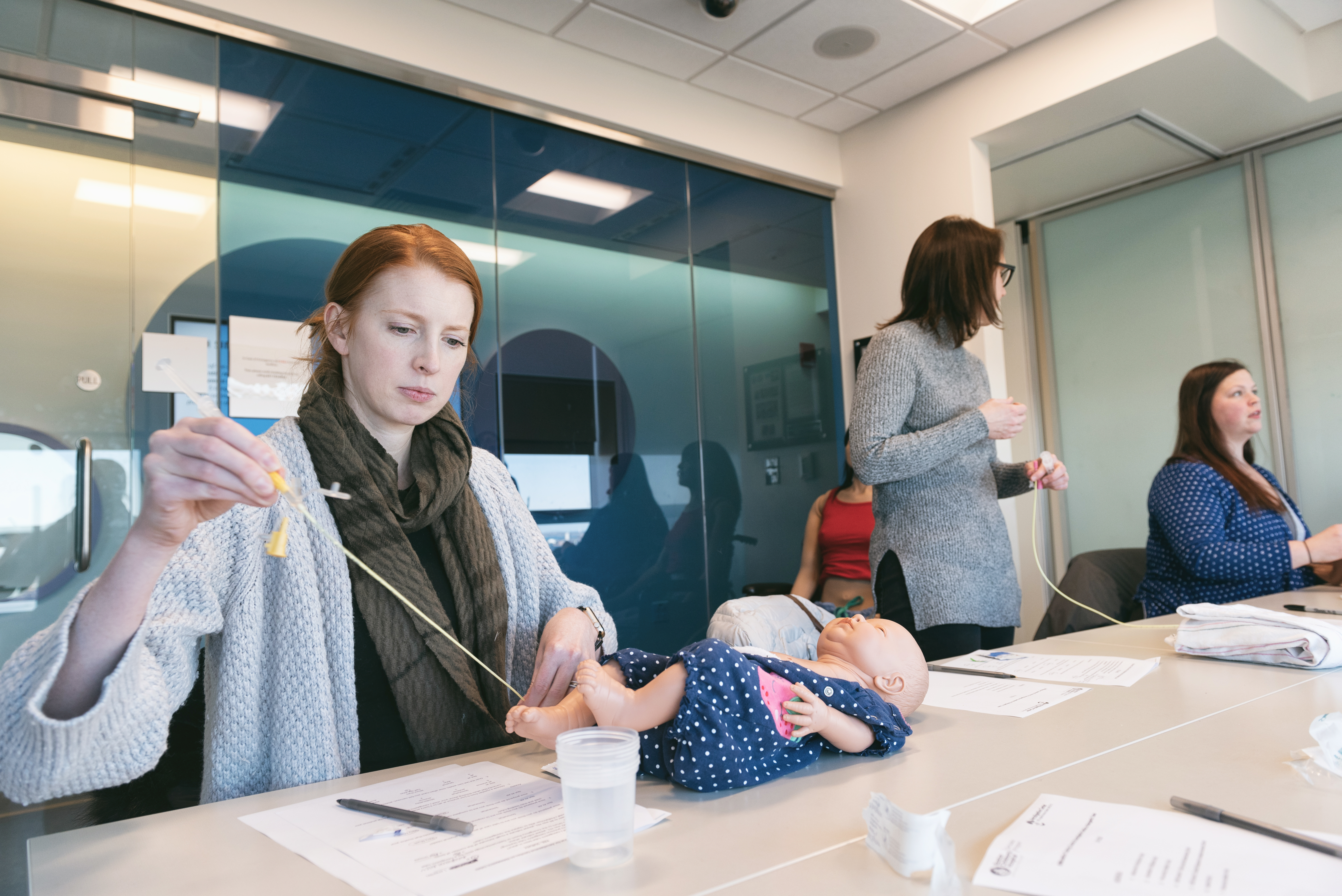 Woman with red hair, green scarf, and grey sweater sits at a table as she practices incisions on a baby doll.