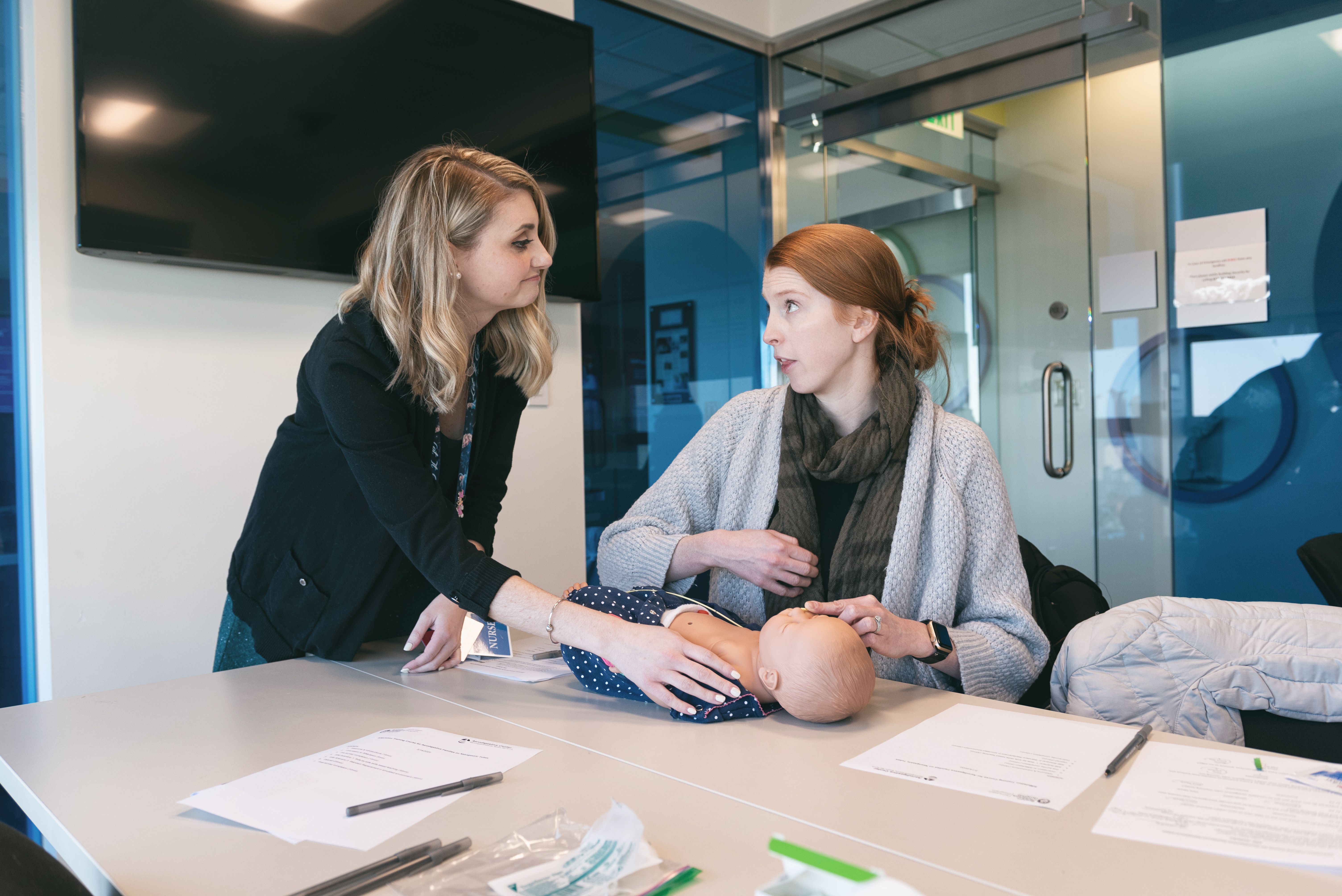 A blond woman and a red-haired woman practice a medical procedure on a baby manikin.