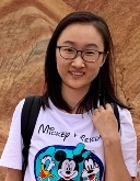 A woman, Zhuo Yang, wears a graphic shirt and glasses in front of a desert rock as she plays with her hair and smiles at the camera.