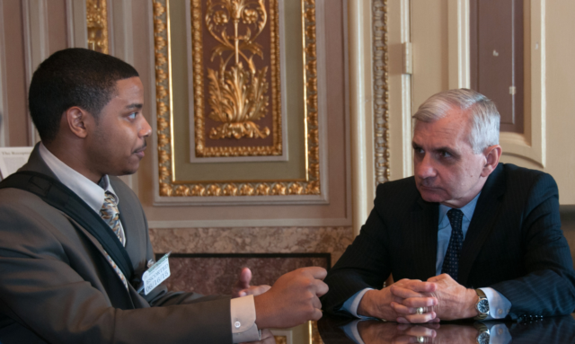 Two well-dressed men sit at a desk and discuss issues