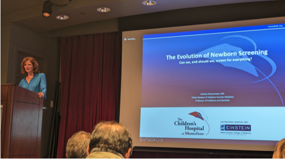 Melissa Wasstein stands at a podium with a slideshow presentation behind her with the title screen reading the evolution of newborn screening and the subtitle: can we, and should we, screen for everything?