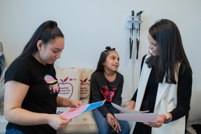 Two women standing and a child sitting, in front of a paper grocery bag of food.