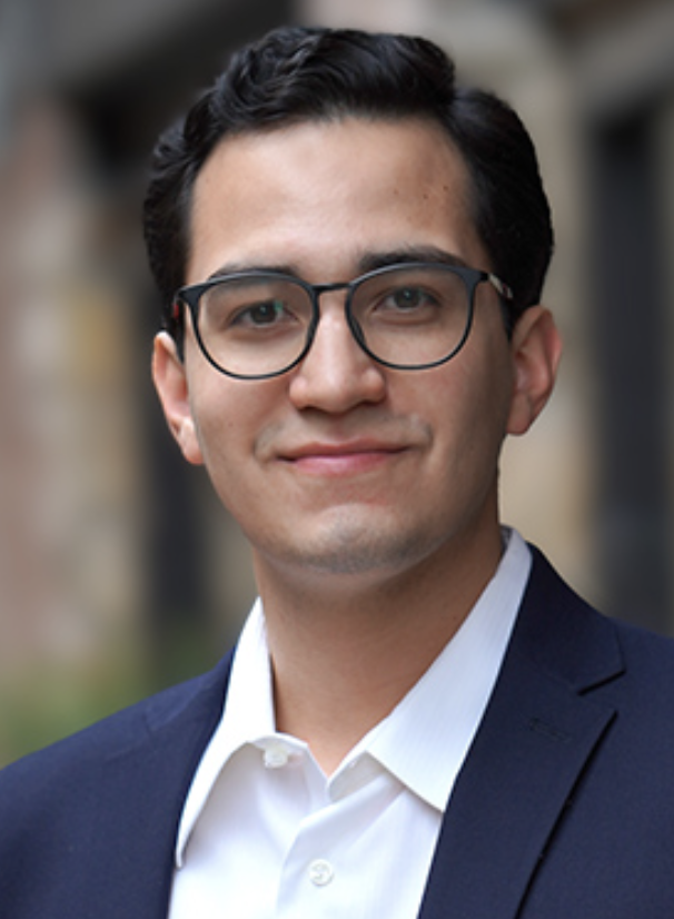 A man, Joaquin Moreno, with dark hair and glasses wears a blue suit for his headshot for the pain and studies lab team page.