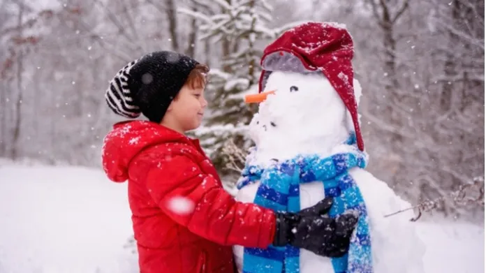 Tweenage boy in winter jacket and hat embraces snowman wearing hat, scarf and carrot as a nose