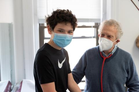 boy wearing mask smiling at the camera while a pediatrician looks on