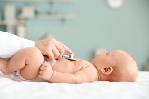 baby on table with a hand holding a stethoscope on his chest