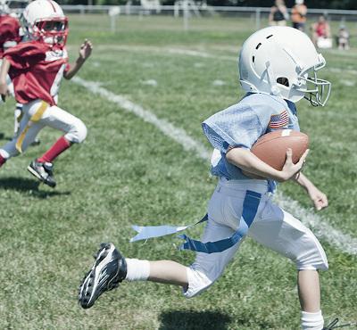 child in football pads and helmet running down field with football