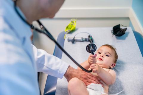 a cute baby holding onto a stethoscope with a doctor leaning over him