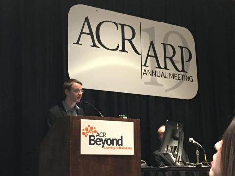 Pierre, a blond man with glasses, stands at the podium at the ACR annual meeting.