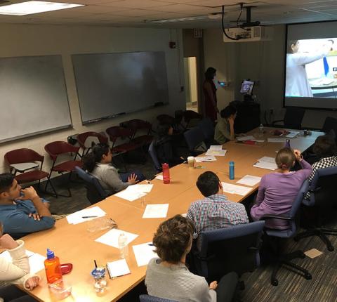 A group of people around a table watch a powerpoint presentation.