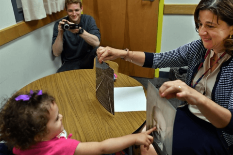 A woman shows a toddler two images, a railroad and a baby's face, and the toddler points to the baby's face.