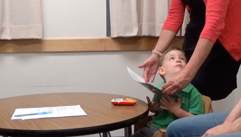 A woman, out of frame, stands behind a toddler boy who looks back at her from his chair at a table.