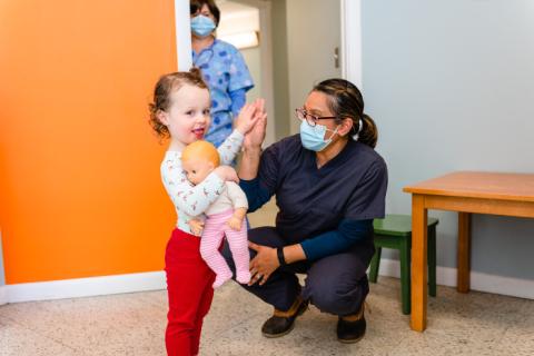 a child holding a doll high fiving a doctor squatting down to her eye level and wearing a mask