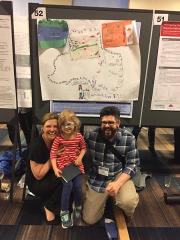 A family holds their child on top of their knee as they pose in front of a hand-drawn map.