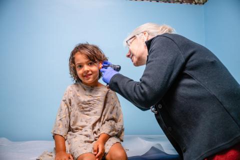 a young girl looking at the camera while a female pediatrician bends over and looks in her ear