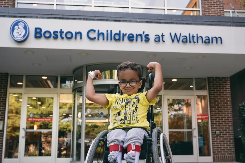 Boy in wheelchair flexes arm muscles in front of sign at entrance to Boston Children's Waltham