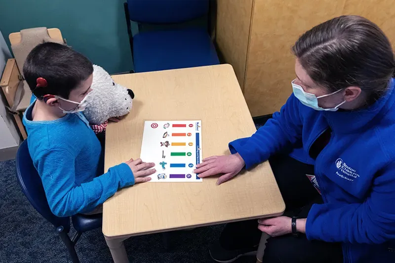 Clinician on right wearing jacket and face mask works with young boy (seated at left) on speech